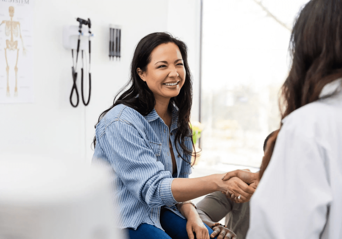 A woman smiling warmly while shaking hands with another person in a bright room.